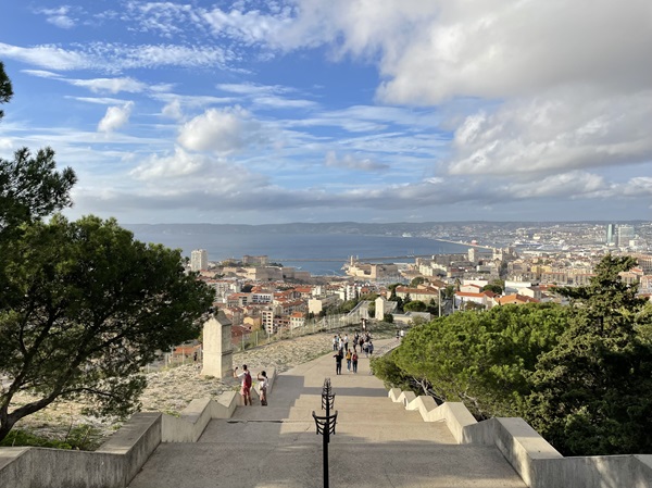 View from the top of a staircase going down to the city of Marseille with water and a blue sky in the background.