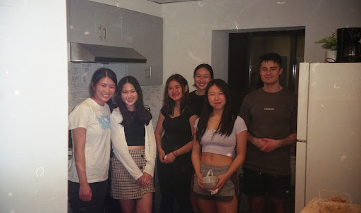 Six students stand in a kitchen with white walls, grey cabinets behind them and a white refrigerator to their right