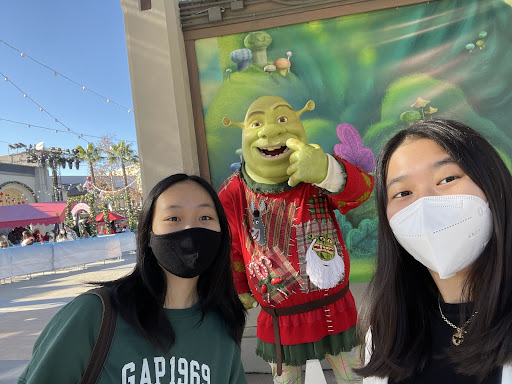 Selfie of two woman and Shrek character, in front of a fantasy backdrop, with an outdoor restaurant visible on the left