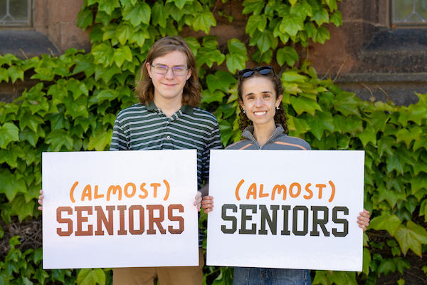 two students holding placards reading "almost seniors" in front of ivy-covered building