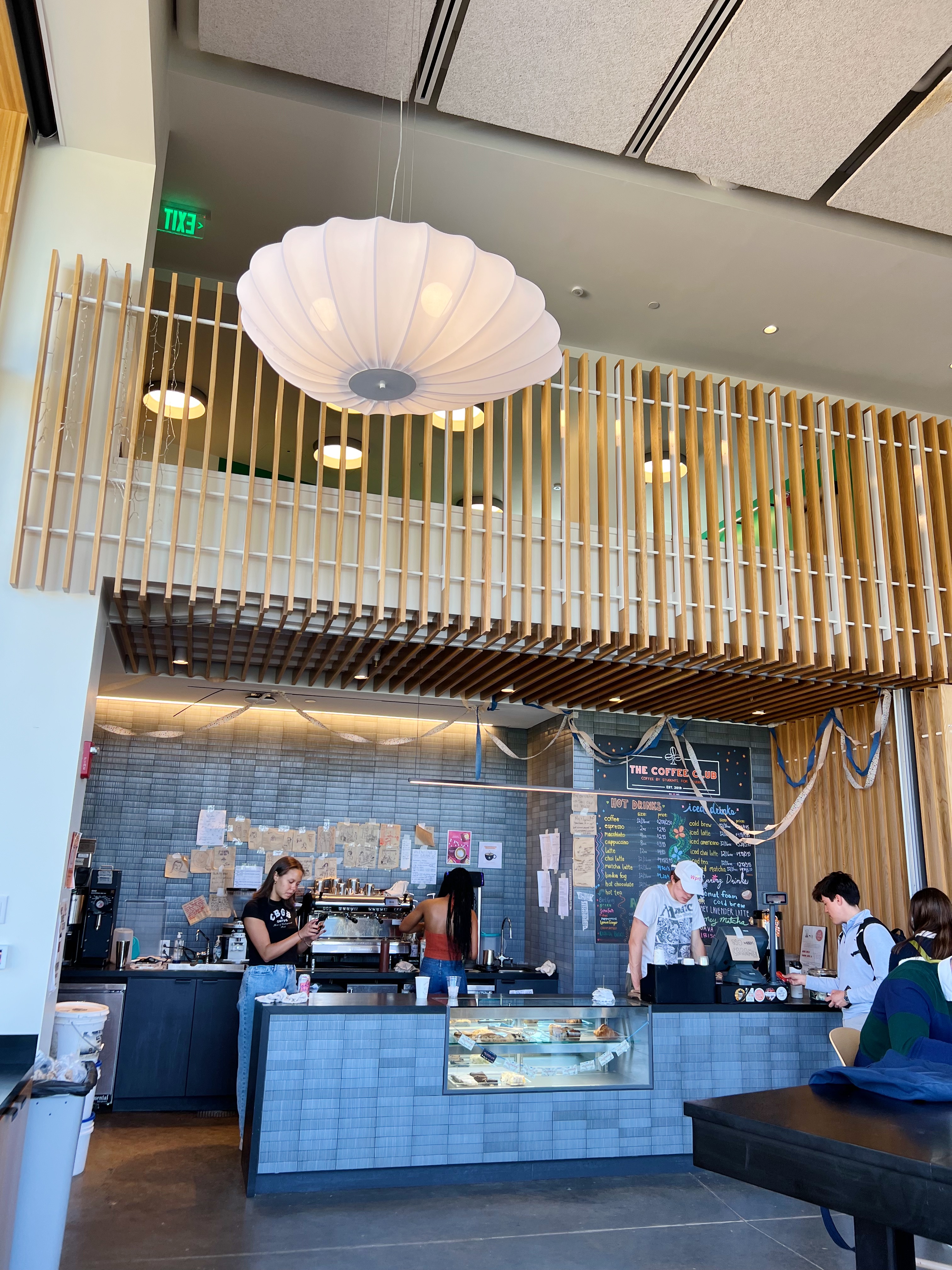 Coffee shop counter with loft above it, light wood railing and paper lantern chandelier