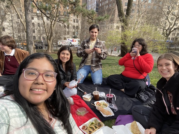 Students sitting on a picnic blanket in the park eating food. 