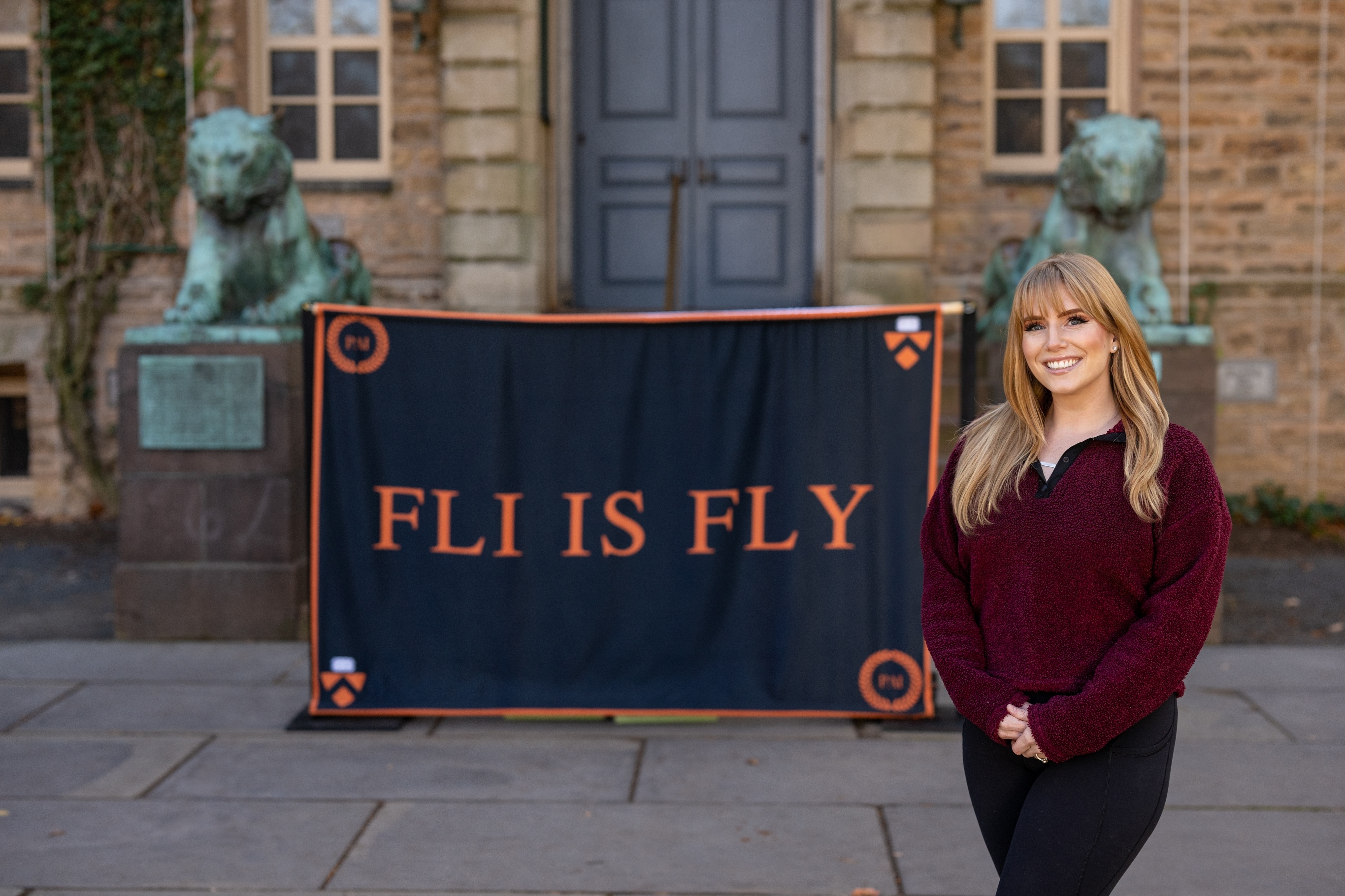 Woman stand in front of banner reading "FLI IS FLY," in front of stone building with two tiger sculptures
