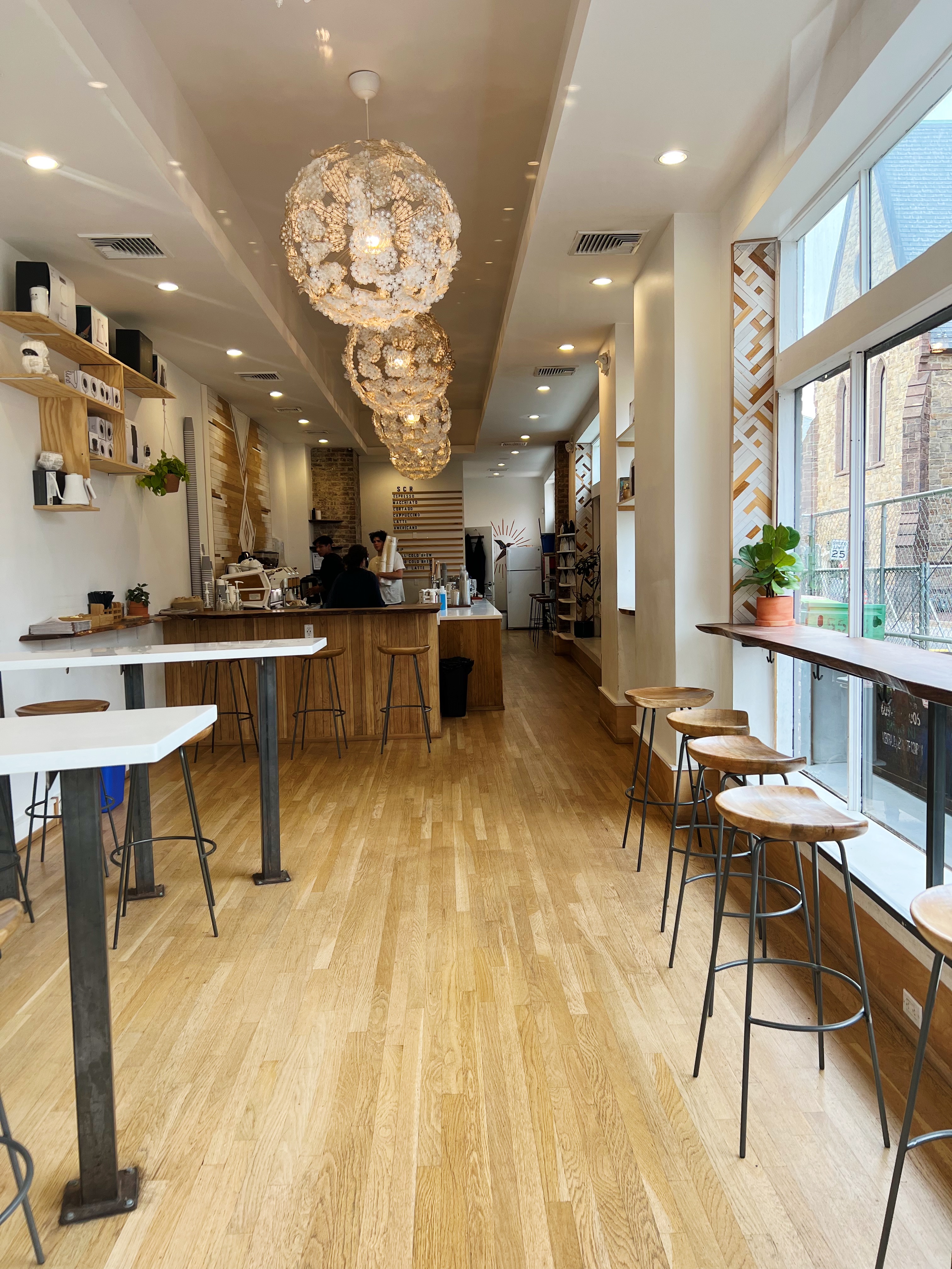 Interior of a coffee shop with light wood floor, high top tables, a bar with stools against the window and floral chandeliers