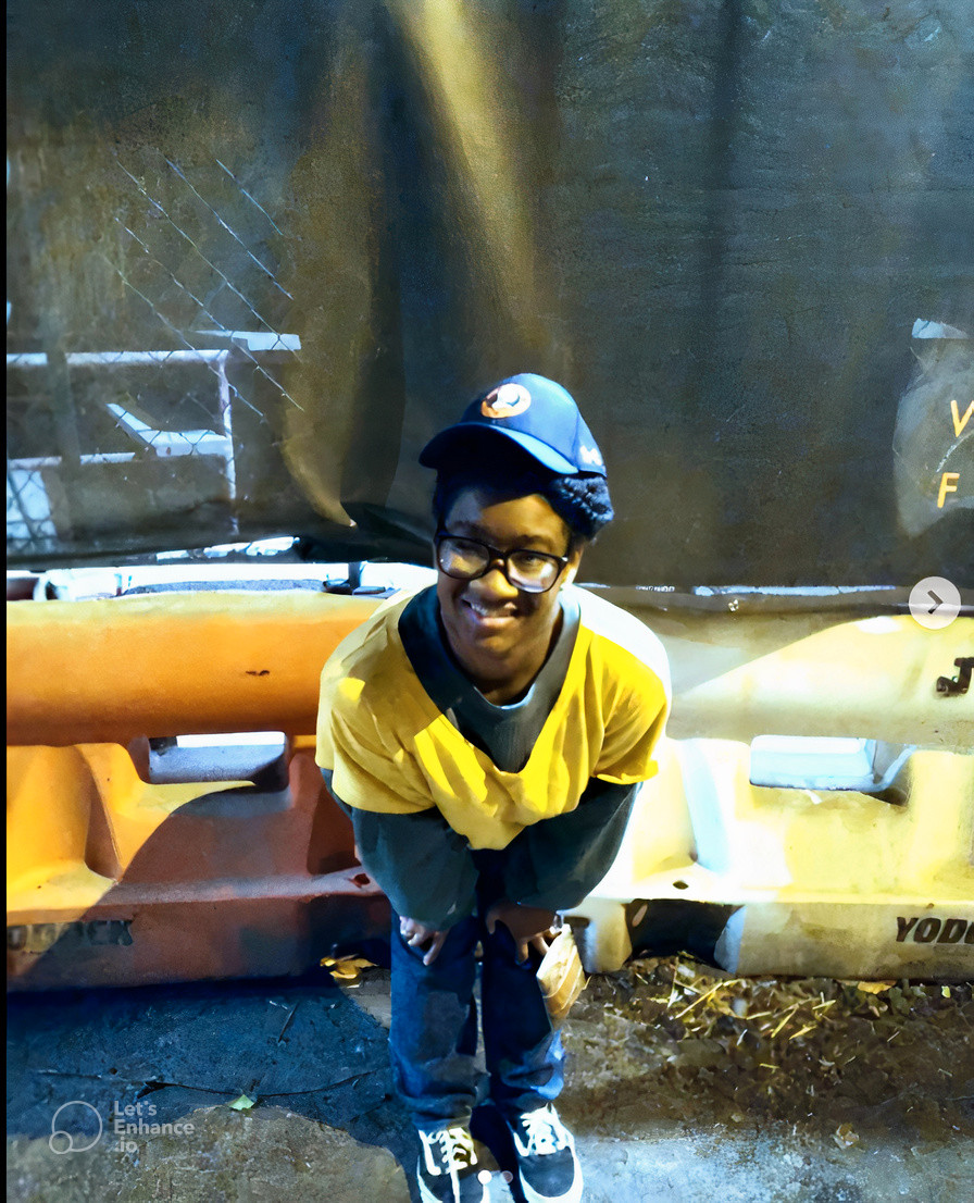Avery is standing in front of the Princeton Builds Pathways construction post, wearing yellow and green shirts and a navy blue Princeton University cap.