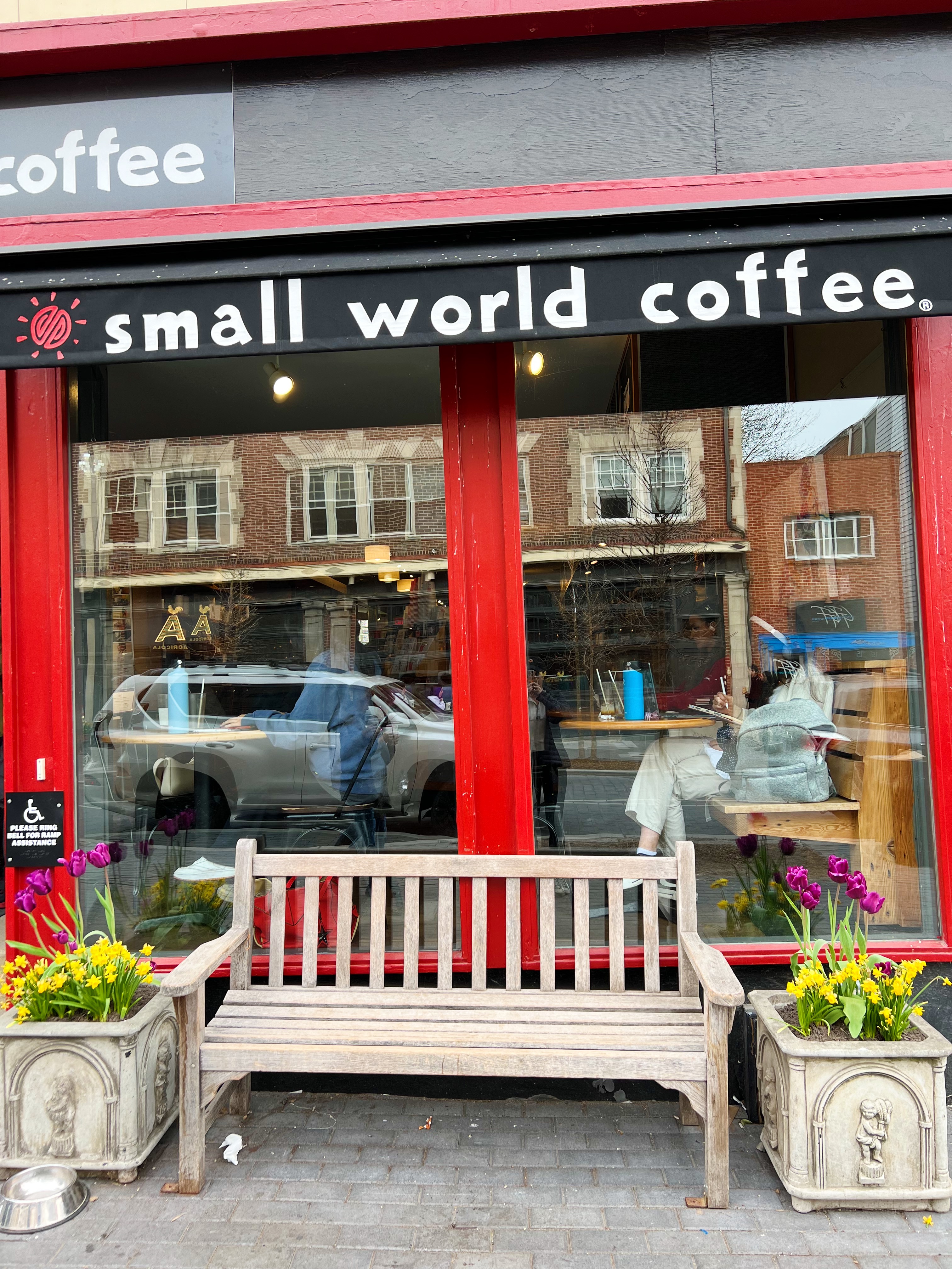 Exterior of Small World Coffee with a bench, floral planters, red lacquer paint and "Small World Coffee" awning