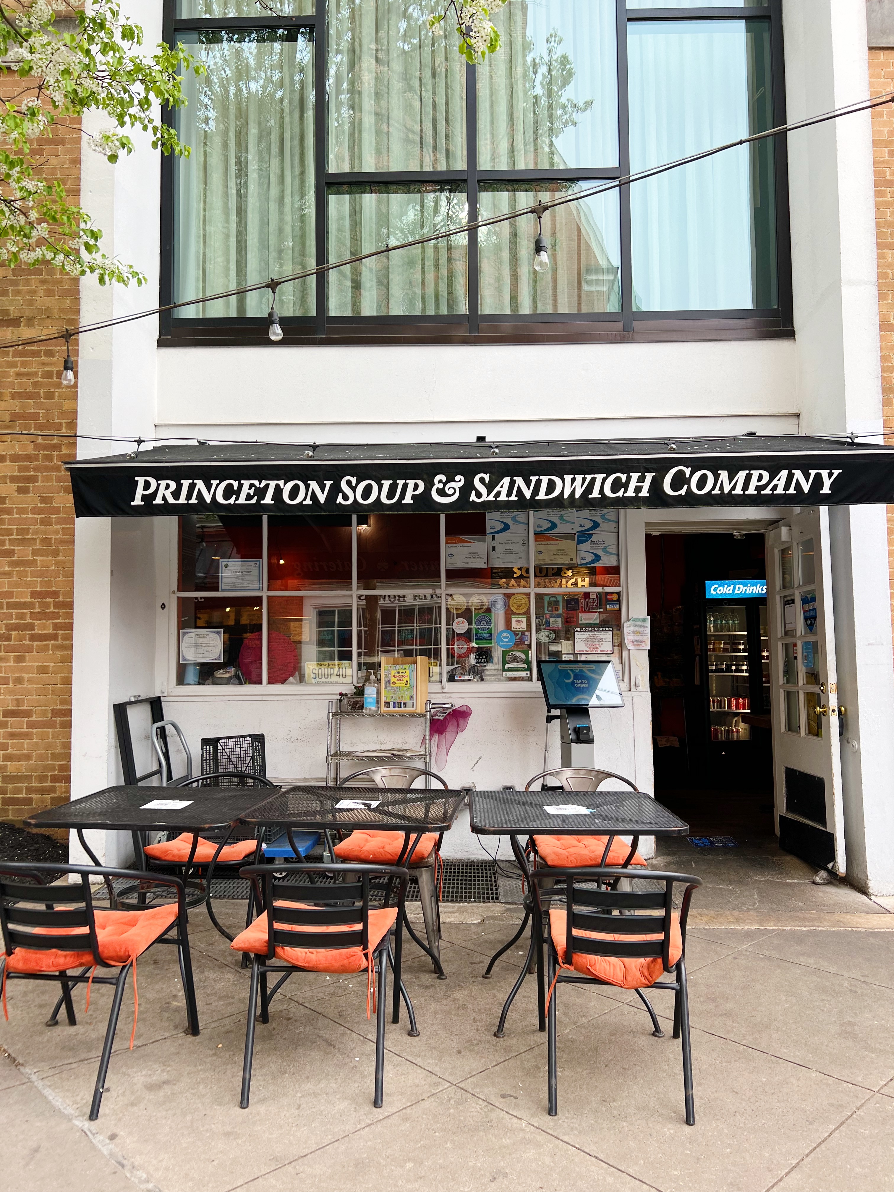 Exterior of Princeton Soup & Sandwich, white building with black trim, black "Princeton Soup & Sandwich" awning, and black metal chairs and tables with orange seat cushions