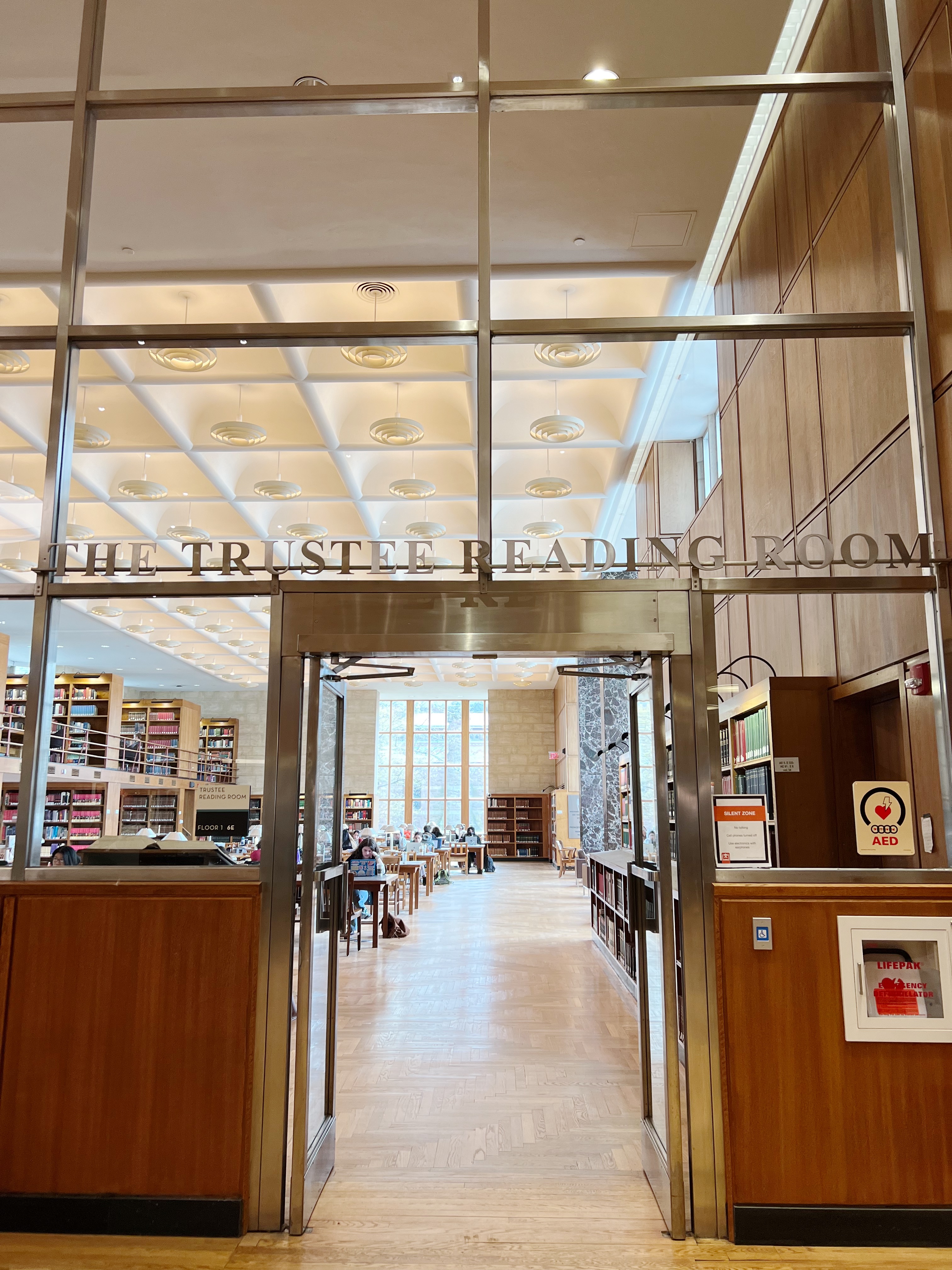 Glass wall with doorway entering a room with tables, letters reading "the trustee reading room" above the door