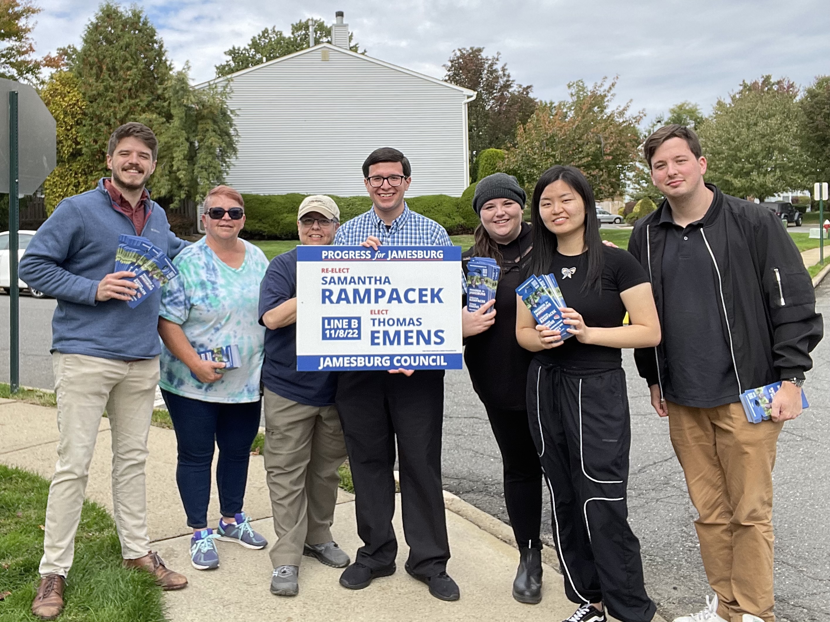 seven people stand on suburban street corner, center person holds campaign sign