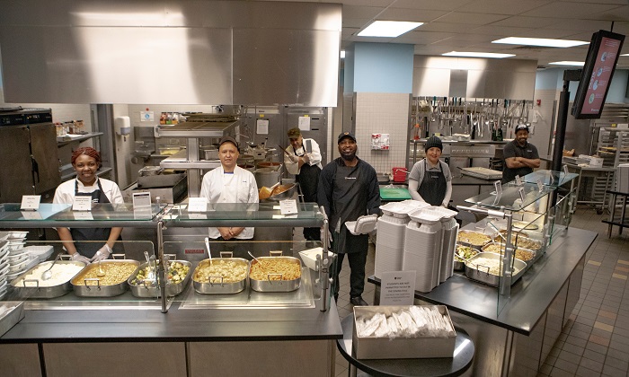 Cinco trabajadores de los comedores del campus están detrás de la cubierta listos para servir comida.