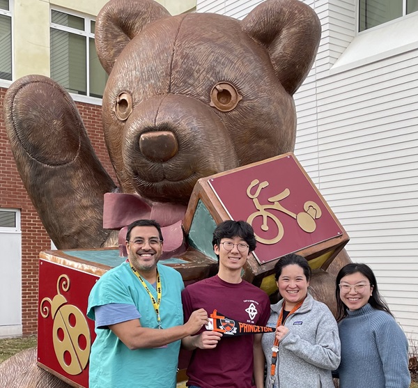 Myself, the other Princetern and our alumni hosts holding a Princeton pennant in front of a giant teddy bear structure holding play cubes