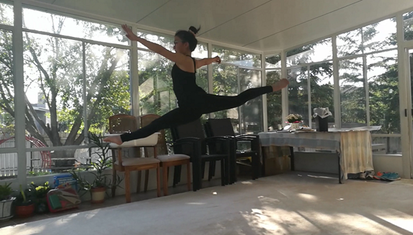 Me, a girl wearing a black tank top and black leggings, doing a split jump in a sunroom