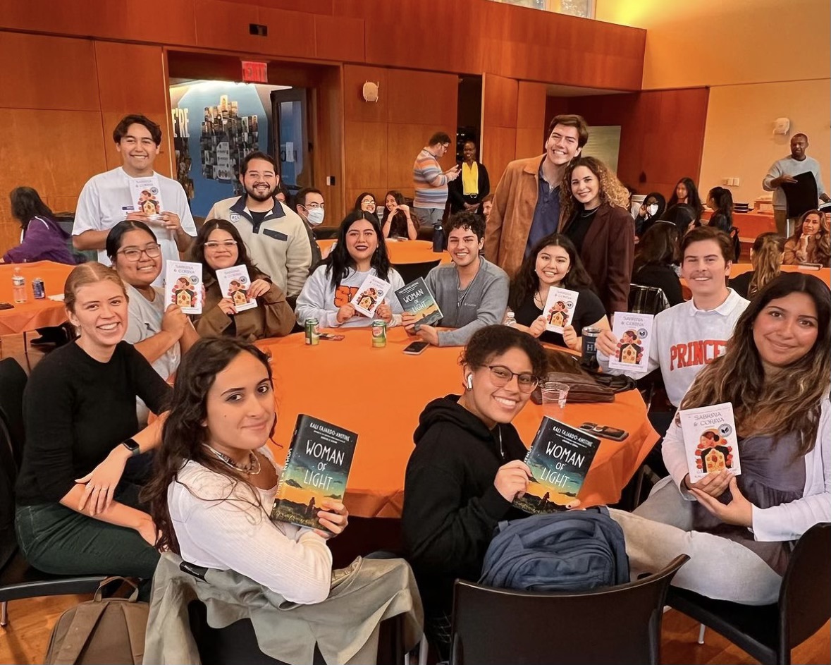 Students sit around a round table with an orange table cloth, holding up books.