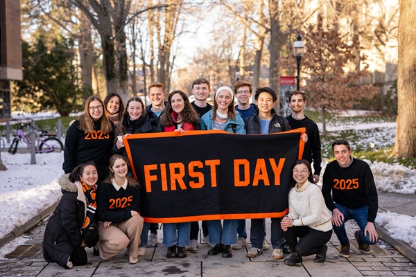 Group of people around a black banner with “First Day” in orange text