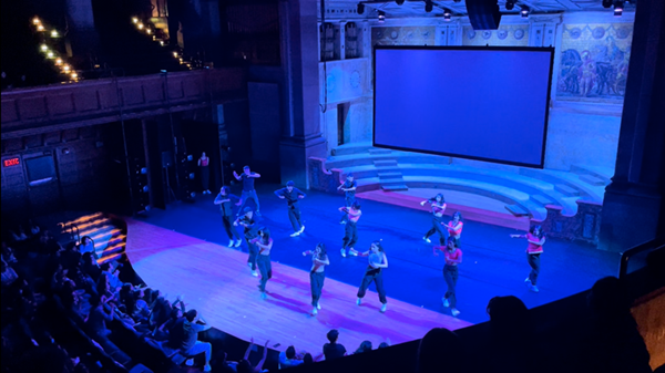 Balcony view of dancers on a large stage under blue light in front of an enthusiastic audience
