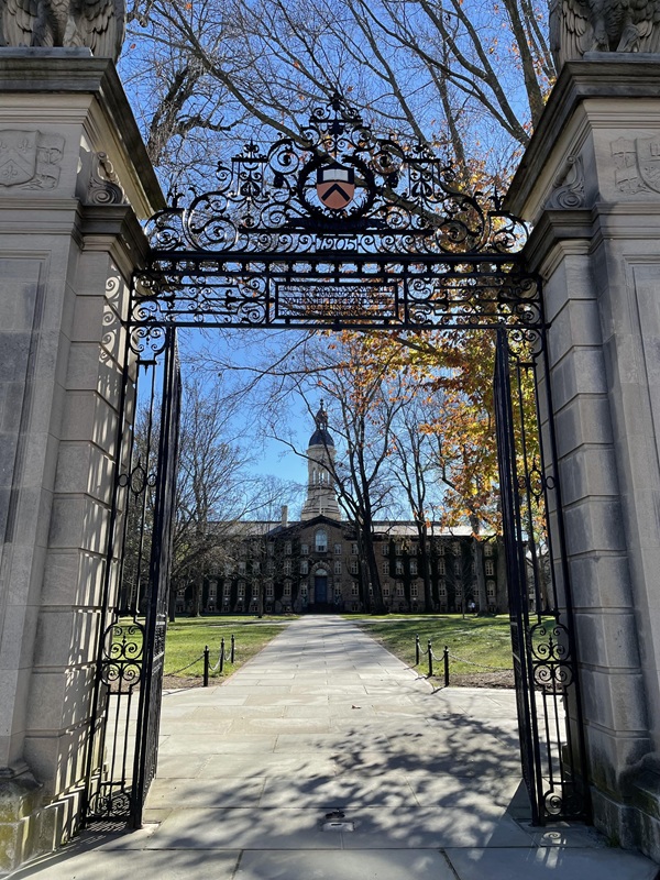 Photo of a wrought-iron gate with stone columns on either side. Nassau Hall, an ivy-covered building with a white cupola, is visible through the gate.