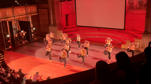 Balcony view of dancers on a large stage under red light in front of an enthusiastic audience