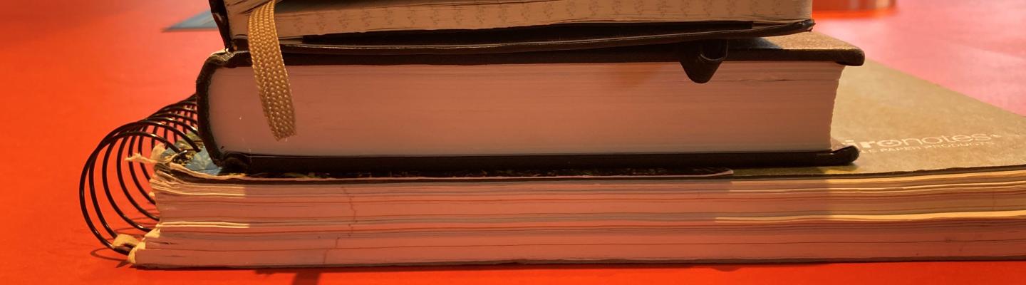 A stack of a large spiral notebook, a black hardcover textbook and a black moleskin notebook on an orange desk at Firestone. In the background, large windows can be seen.