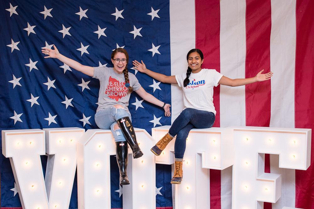 Two students sitting on top of letter that spell Vote with an American flag in the background